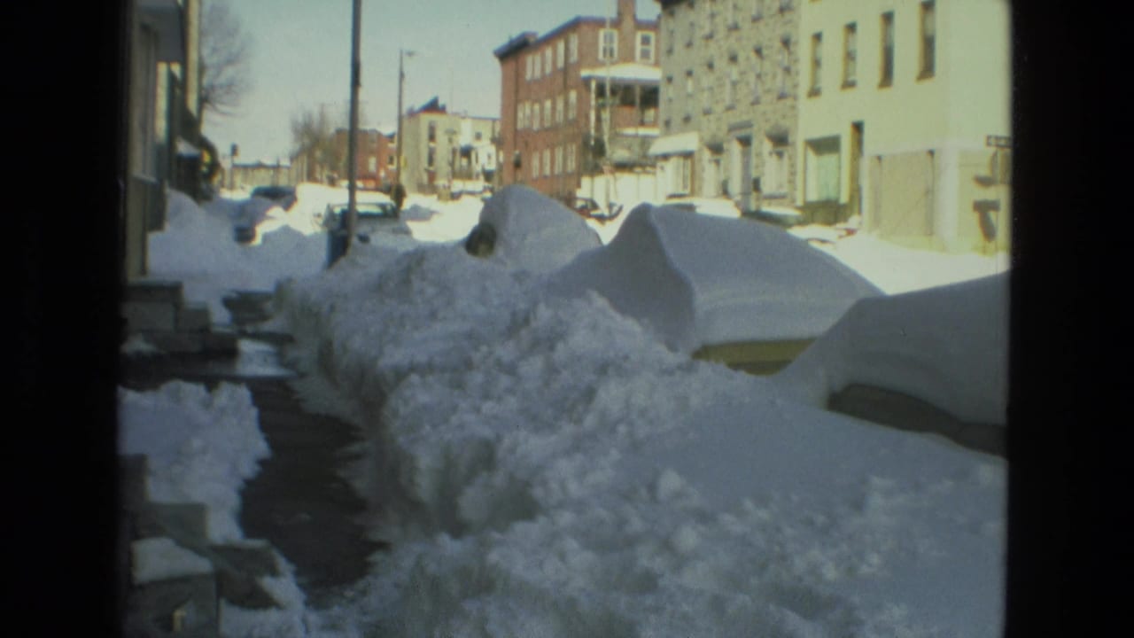 Snow-covered parked cars and sidewalk run past brick residential buildings.