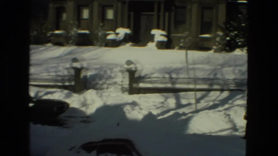 Elevated home-movie view of a Drexel Hill street in 1978, showing snow-covered parked cars, house facades, a fence line, and long winter shadows.