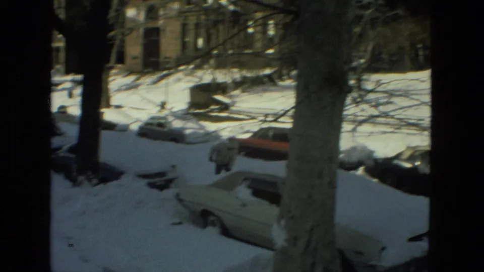 A distant bundled pedestrian walks through a snowy Drexel Hill residential street in 1978, with buried cars, bare trees, and brick buildings in the background.
