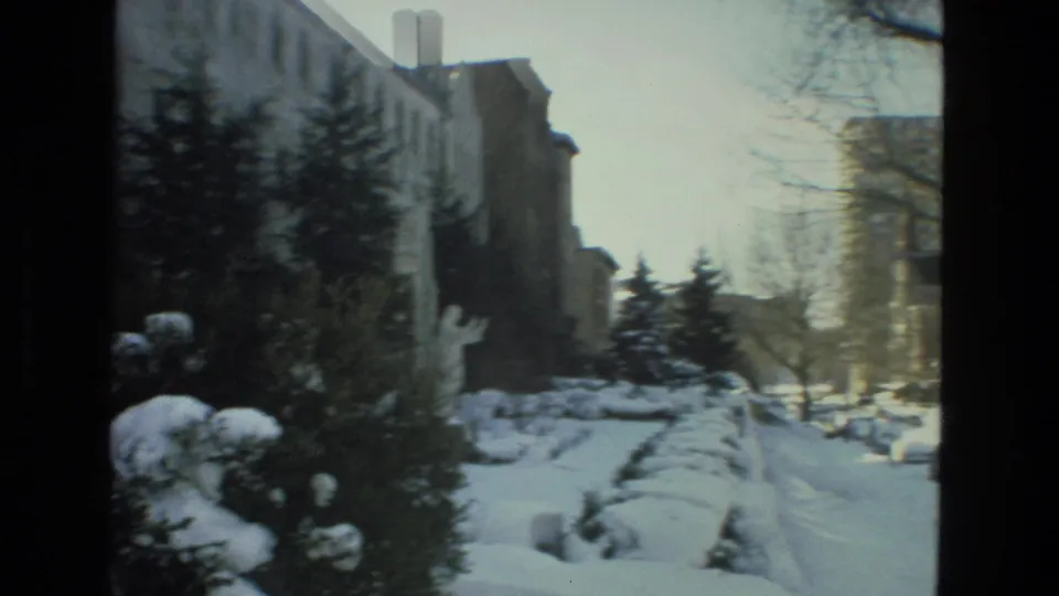 1978 Drexel Hill winter view of snow piled beside a residential building, with evergreens, bare trees, and distant parked cars.