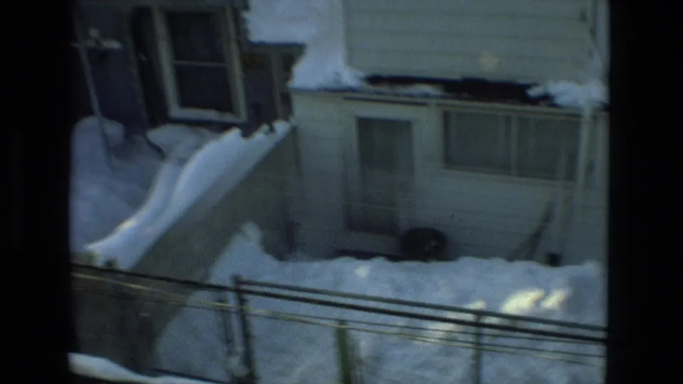 1978 Drexel Hill home-movie frame of a neighboring house exterior with snow piled on the roof, porch, yard, and fence line.