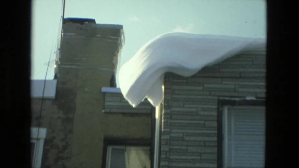 A large overhanging snow drift curls from a residential roof beside a chimney and siding in Drexel Hill, Pennsylvania.