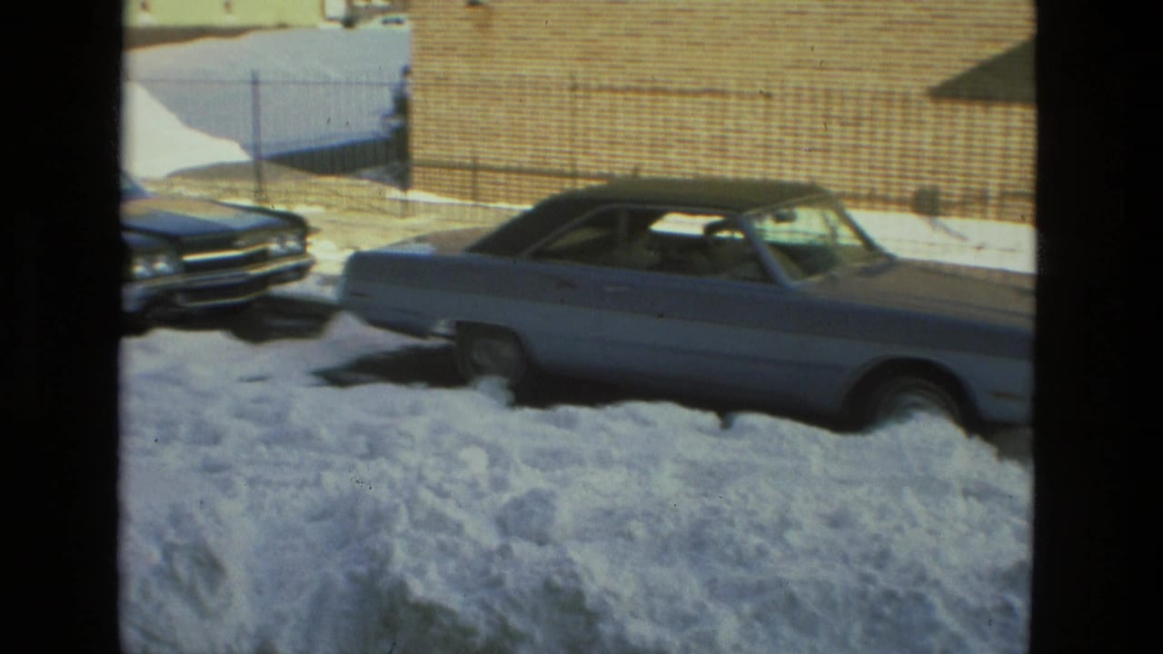 Light-colored vintage coupe parked beside high snowbanks near a brick wall.