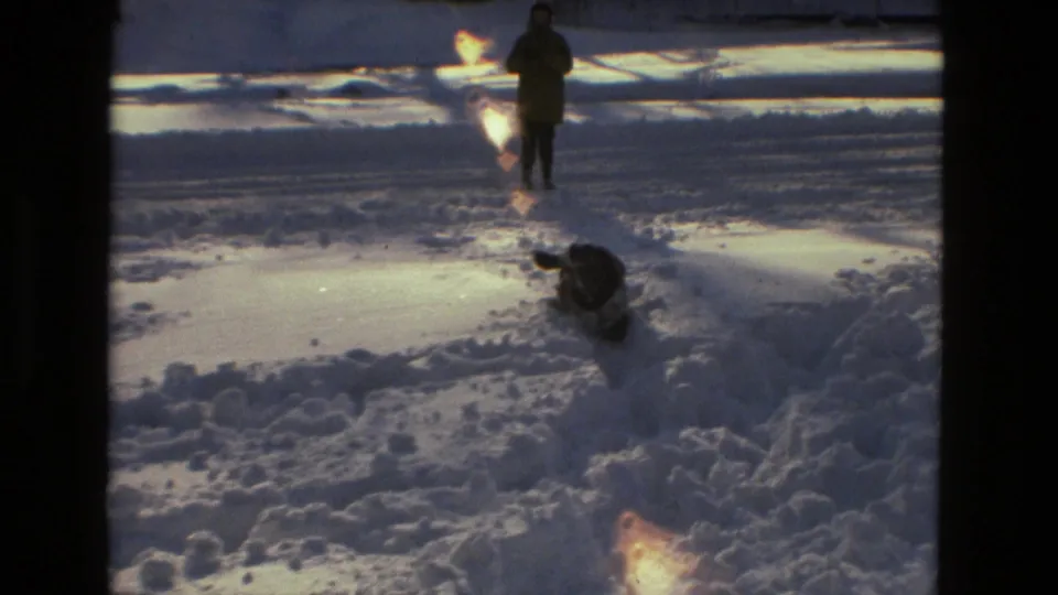 The 1978 home-movie frame shows a distant person on snow and a dark dog moving along a churned path under low winter light.