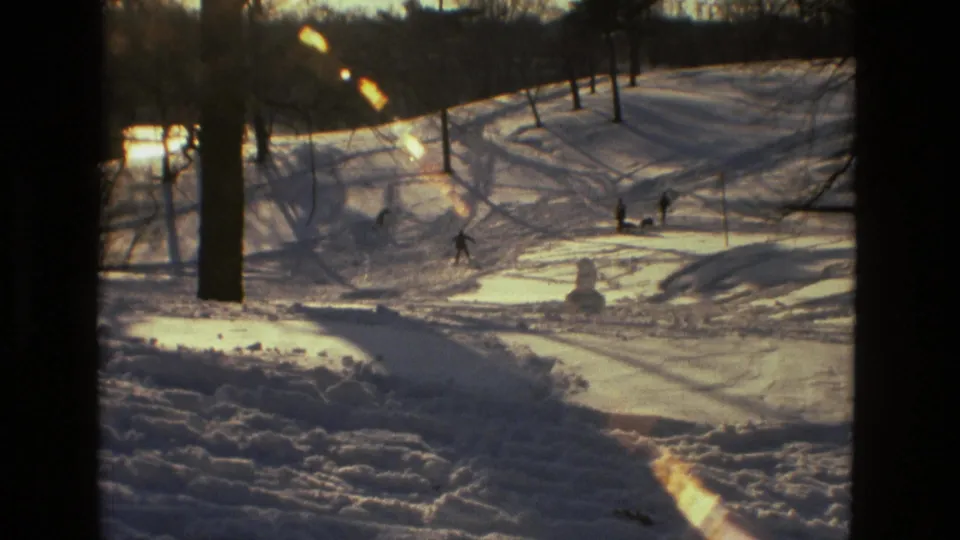 1978 Drexel Hill winter frame with distant people on a snowy slope, bare trees, ski or sled tracks, and a small snowman.