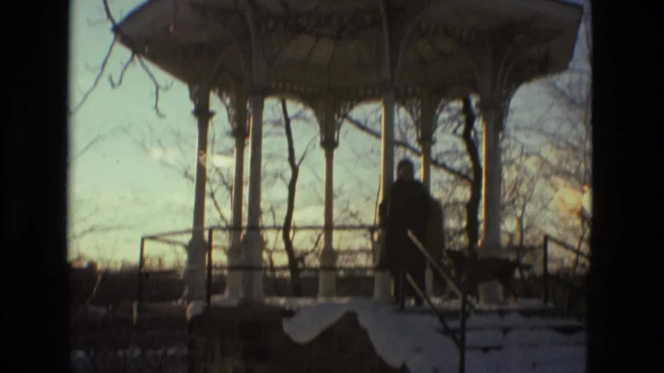 A person in a dark winter coat pauses near a dog by the gazebo, with snow, railings, and bare trees visible in the 1978 Drexel Hill scene.