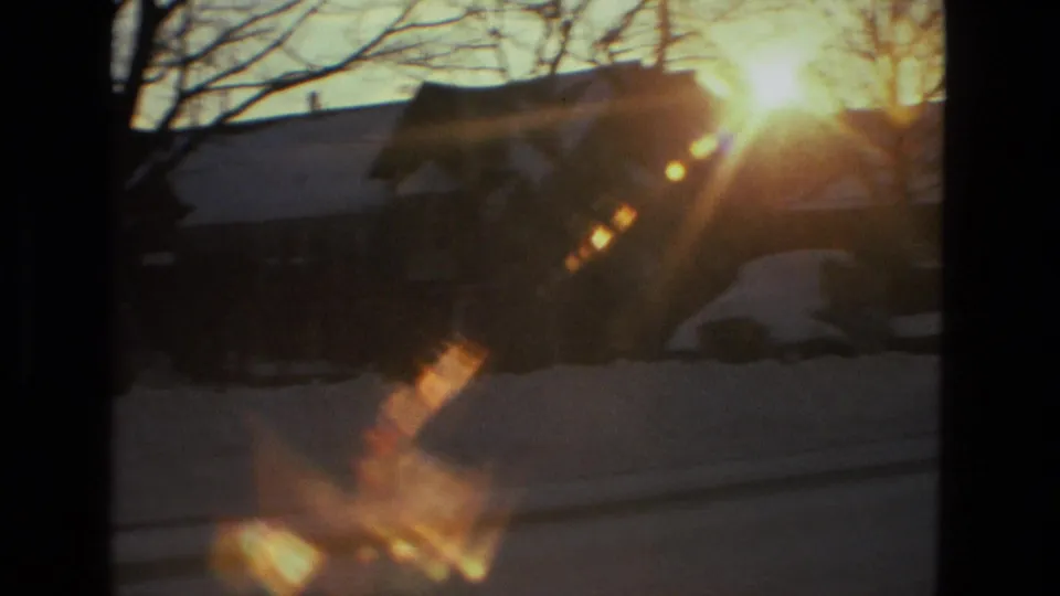 1978 Drexel Hill home-movie frame with bright sun flare over snowbanks, a house roofline, bare branches, and a parked car shape.