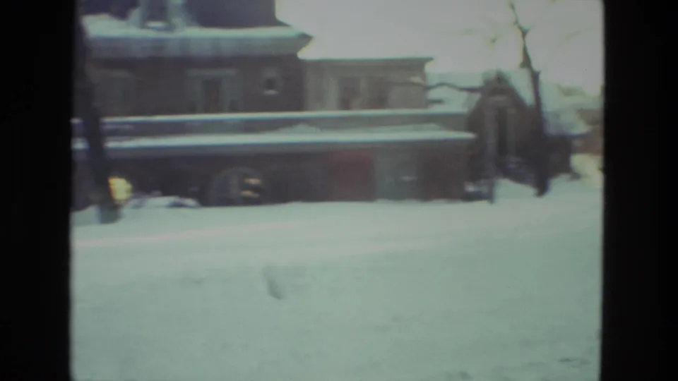 Snow-covered residential exterior in Drexel Hill, Pennsylvania, with porch, roof snow, bare branches, and a distant person near the house.
