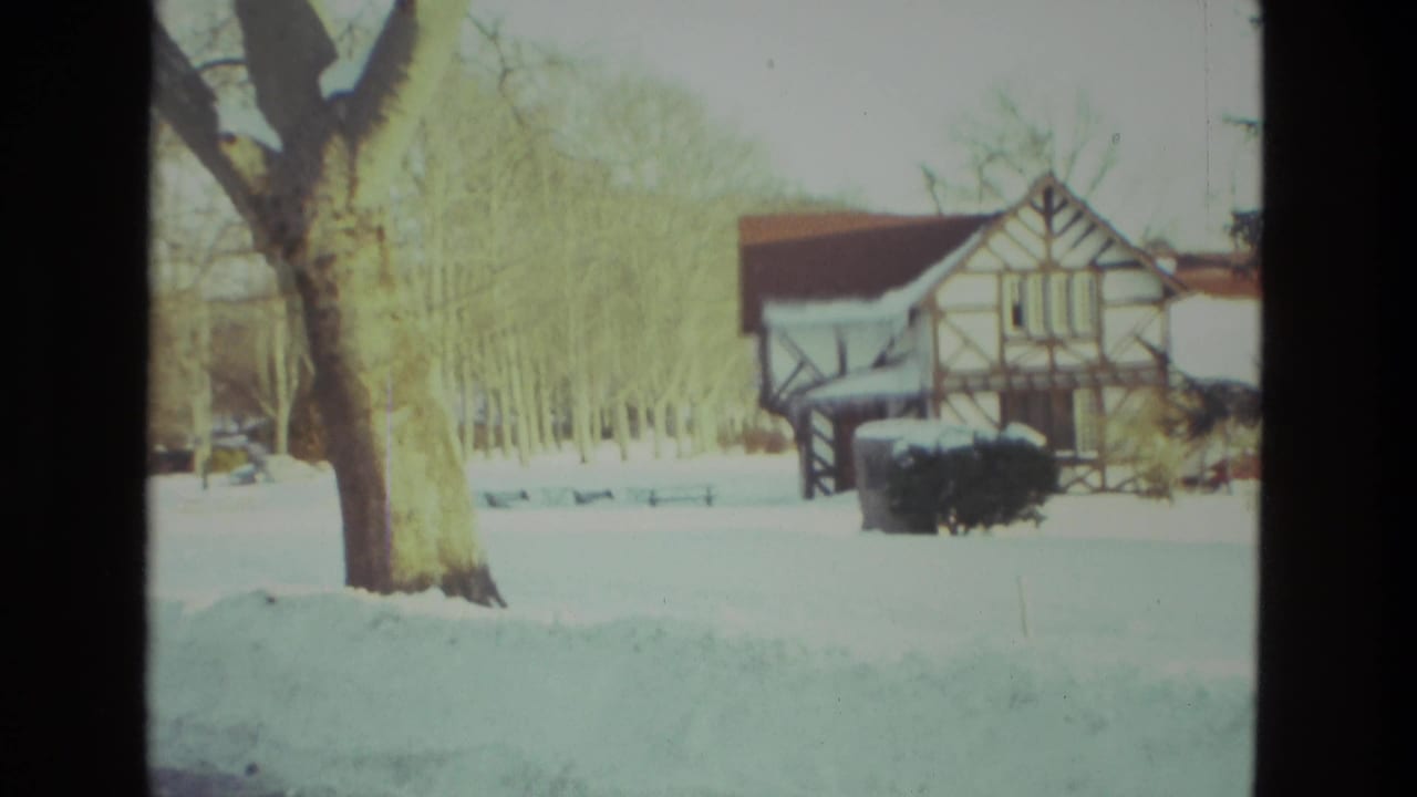 1978 Drexel Hill winter frame with a snow-covered yard, bare trees, and a half-timbered residential building beyond the lawn.