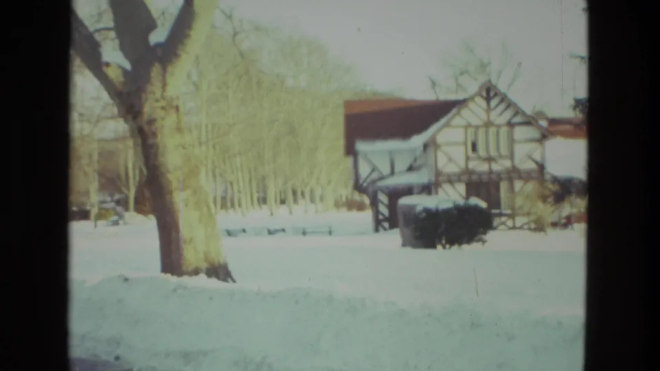 1978 Drexel Hill winter frame with a snow-covered yard, bare trees, and a half-timbered residential building beyond the lawn.