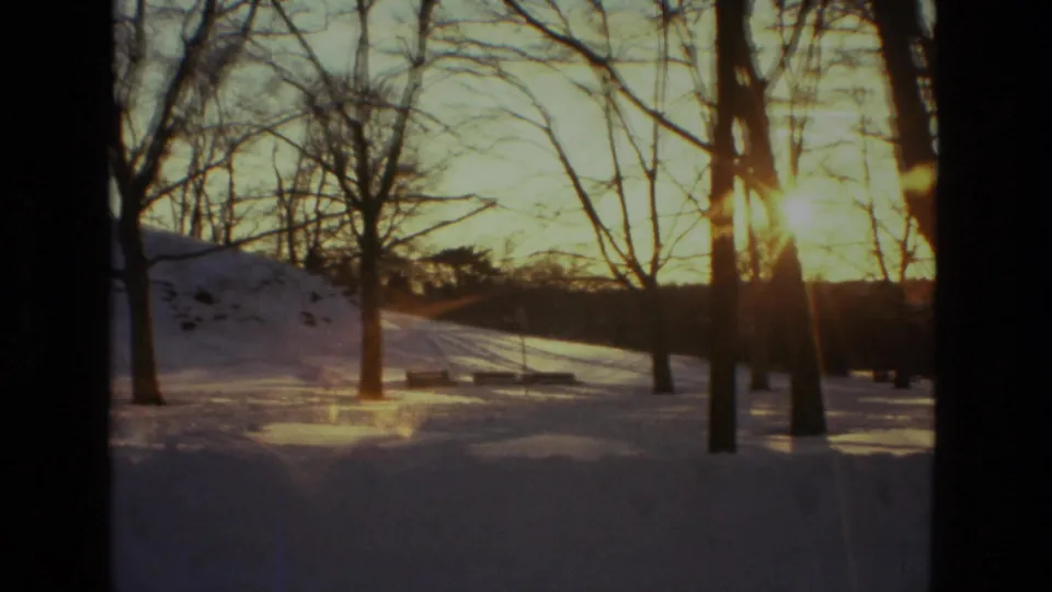 1978 Drexel Hill home-movie frame with warm sunset light through bare trees over snow and a bench-like form in the distance.