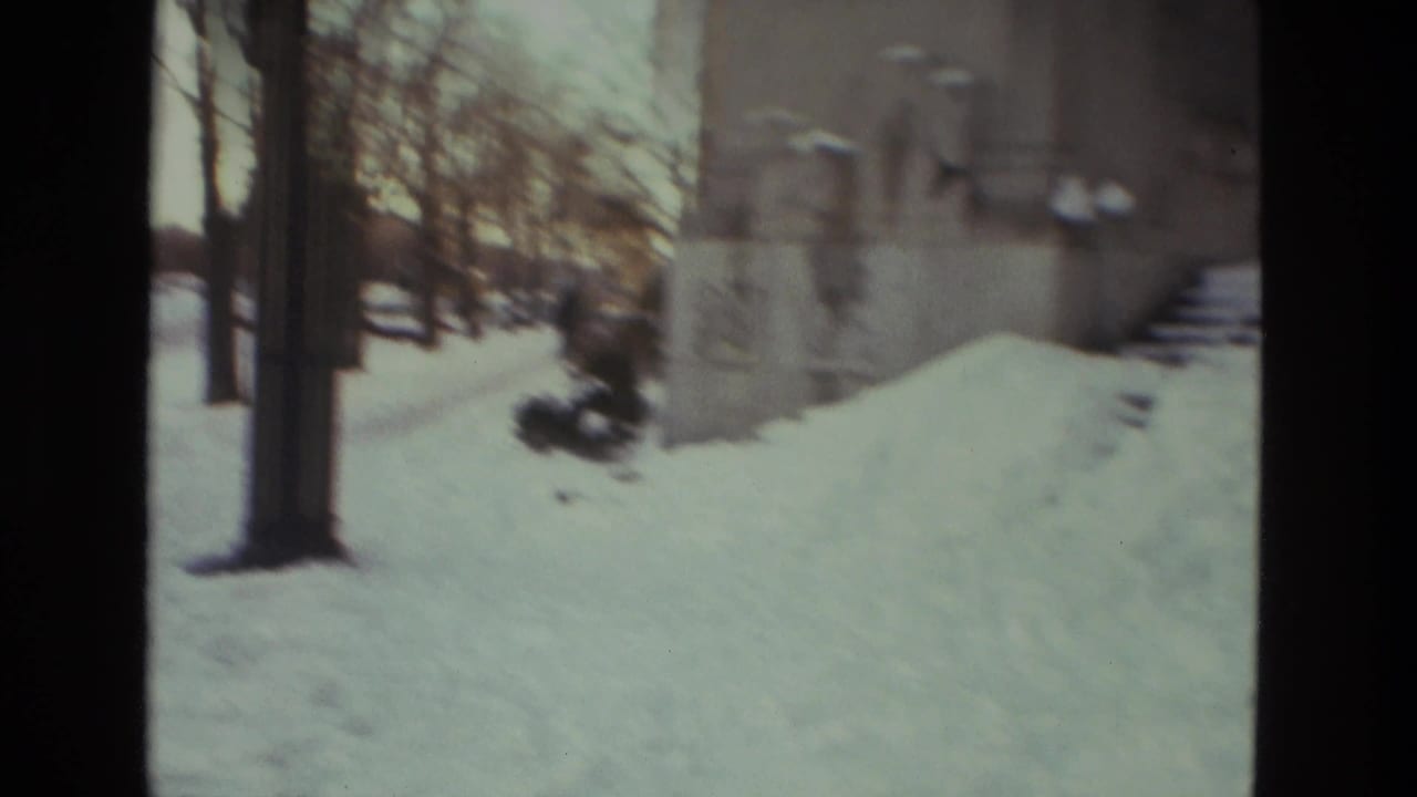 Snow-covered building edge and trees with a dark-coated person moving through deep snow.