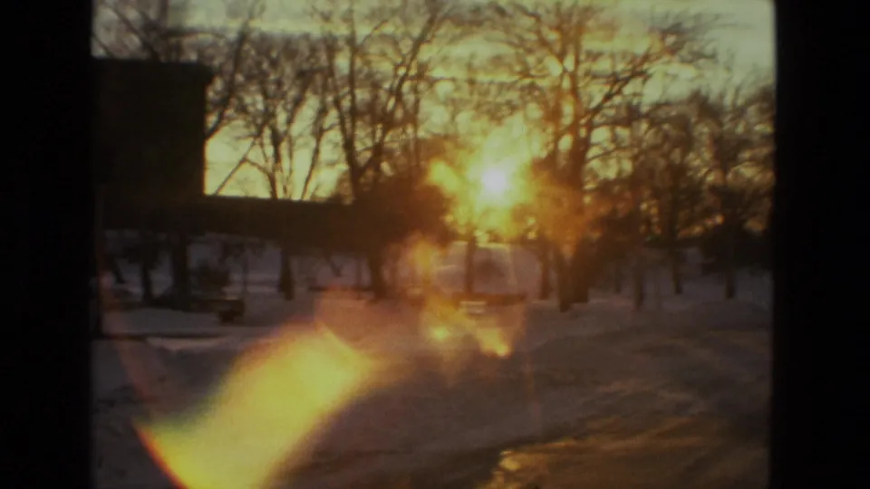Warm sunset glare crosses a 1978 Drexel Hill snowy scene, with bare trees and a dark building edge in faded 8mm color.