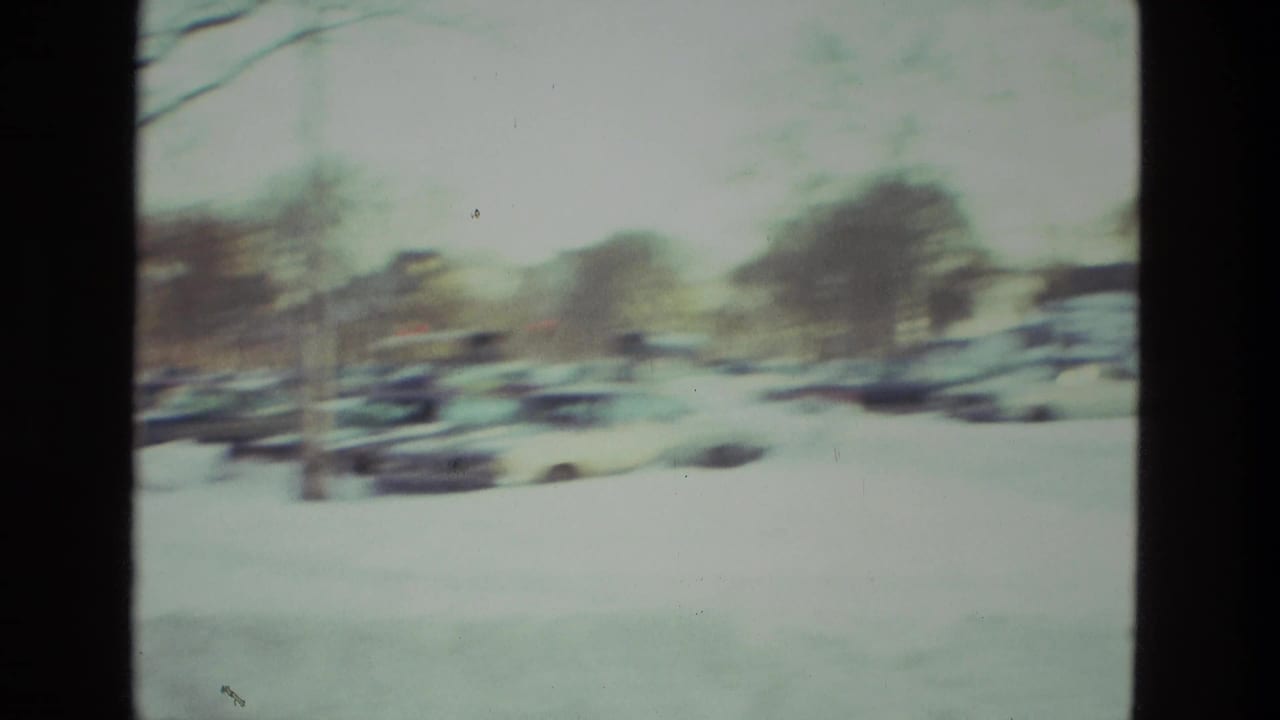 Snowy street with parked cars and bare trees, filmed through a soft handheld 8mm frame.