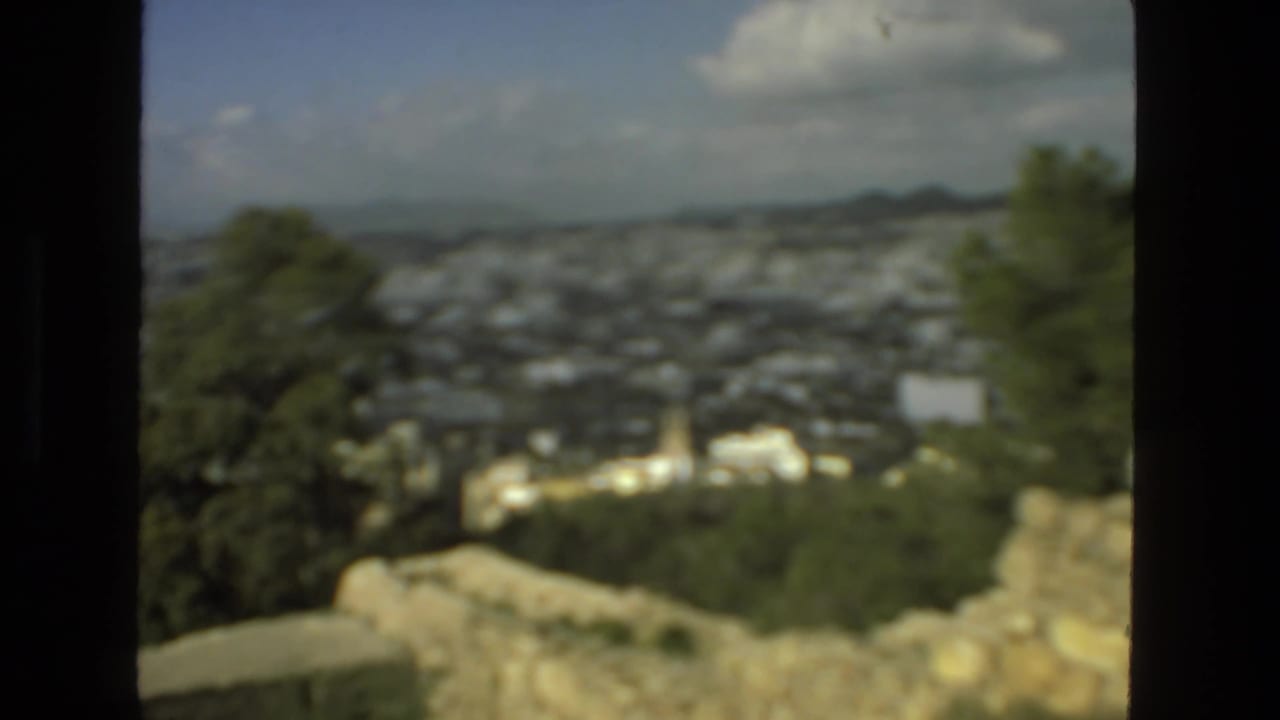 Soft 8mm frame from the Drexel Hill overlook showing trees, a hazy town below, stonework, distant hills, and clouds.