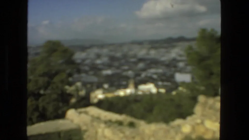 Soft 8mm frame from the Drexel Hill overlook showing trees, a hazy town below, stonework, distant hills, and clouds.