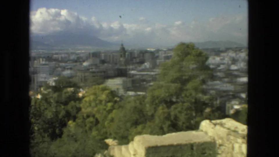 Drexel Hill 1978 home-movie frame showing a town below an elevated viewpoint, with trees, stone foreground, distant hills, and clouded sky.