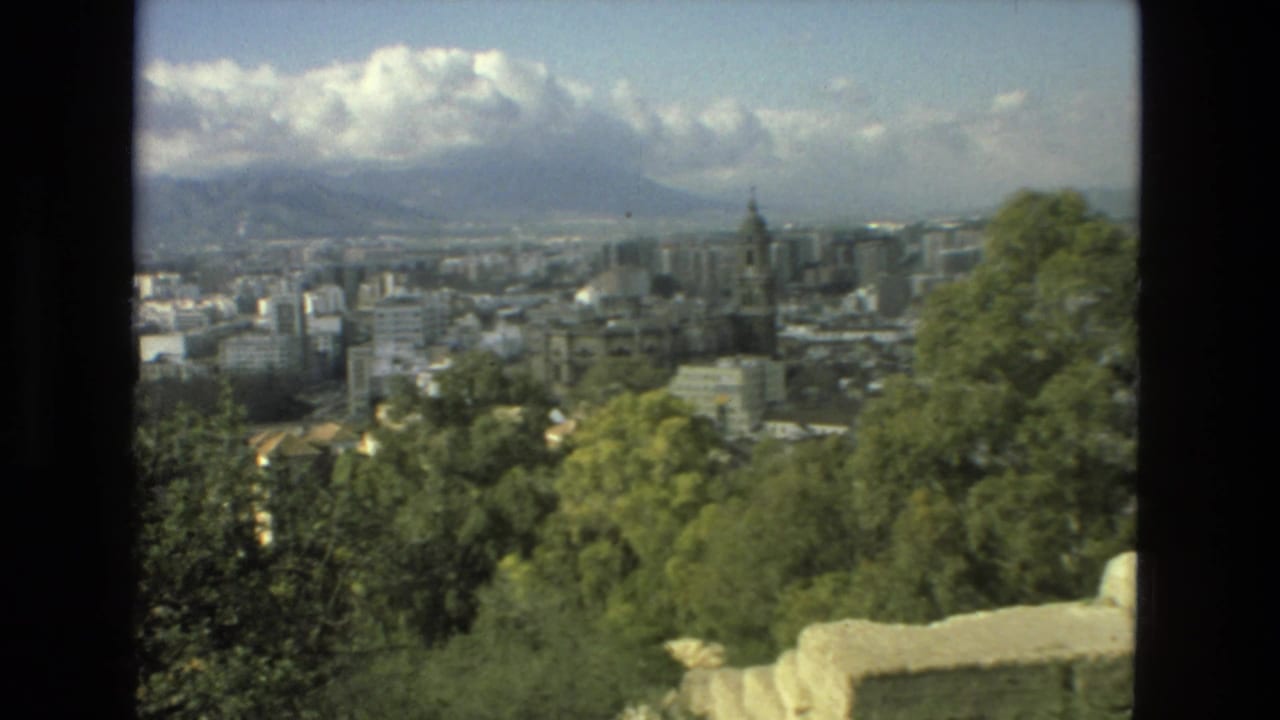 Elevated view over Granada with trees, city buildings, a tower, mountains, and clouds.