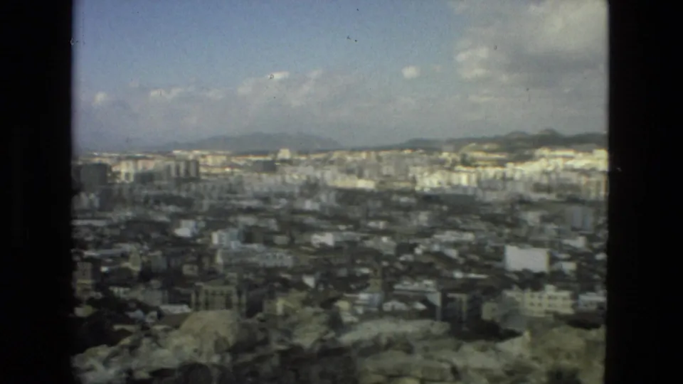 Wide 1978 Granada city view from above, showing dense rooftops, pale apartment blocks, distant hills, and a hazy partly cloudy sky.