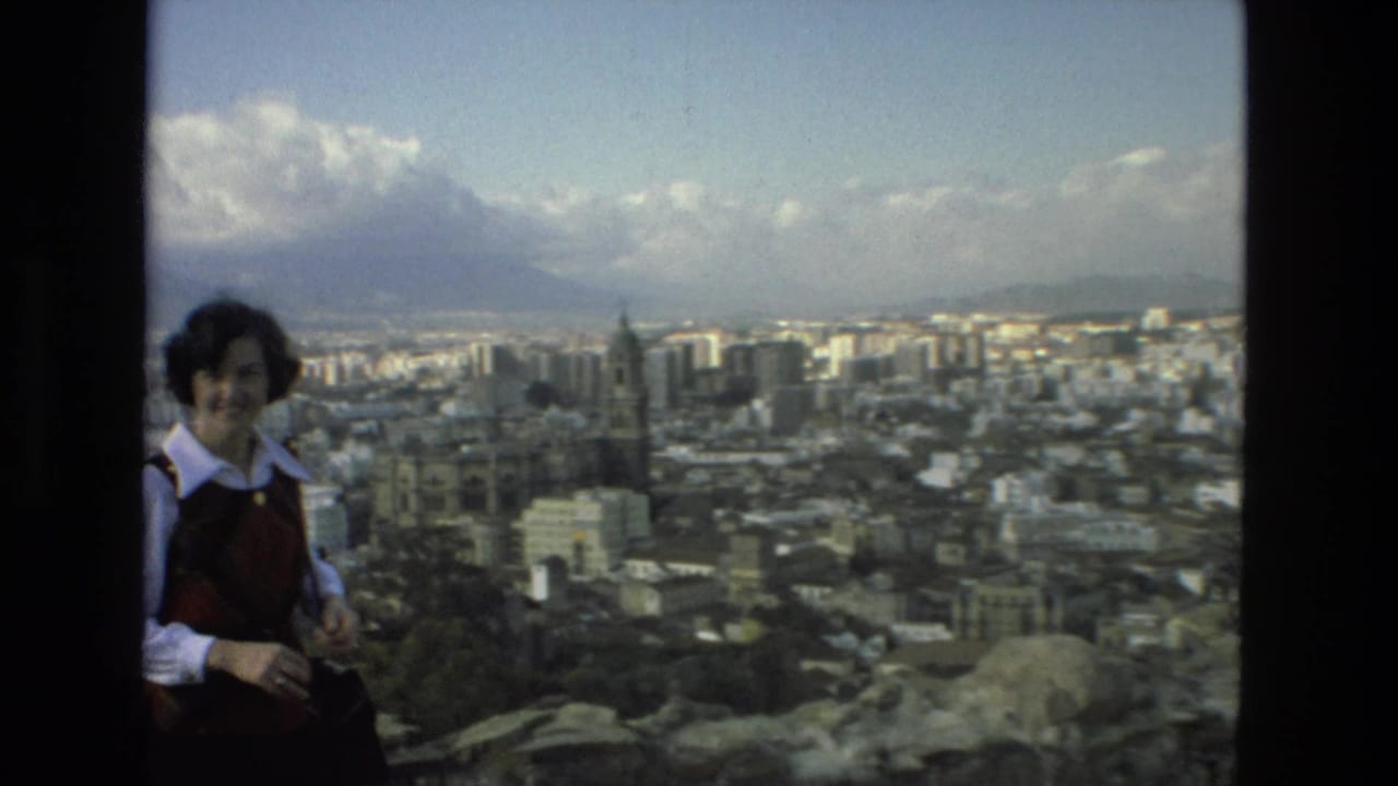 A woman in a white shirt and red vest smiles on a Granada overlook as the 8mm camera frames the city and mountains behind her.