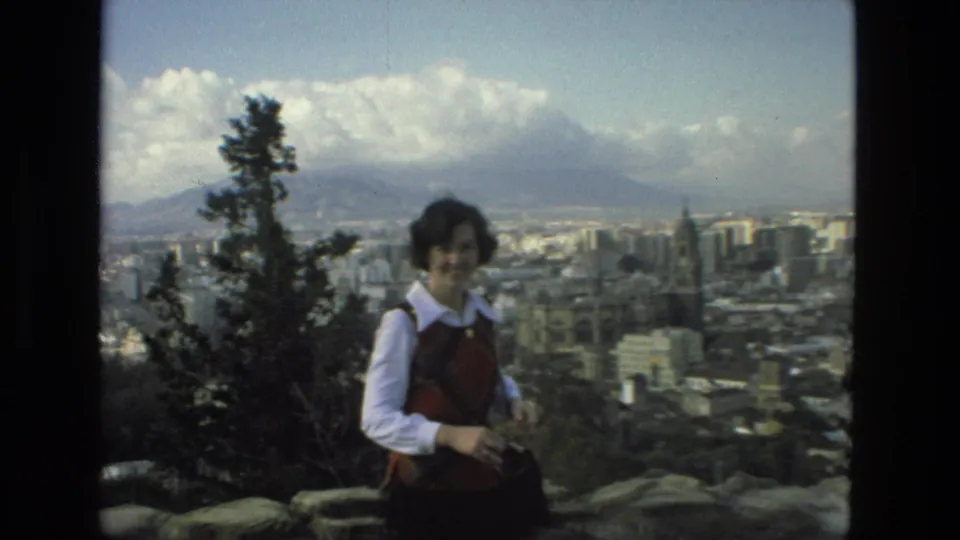 The woman remains on the Granada hilltop overlook, partly framed by a tree, with city blocks and mountains visible behind her.