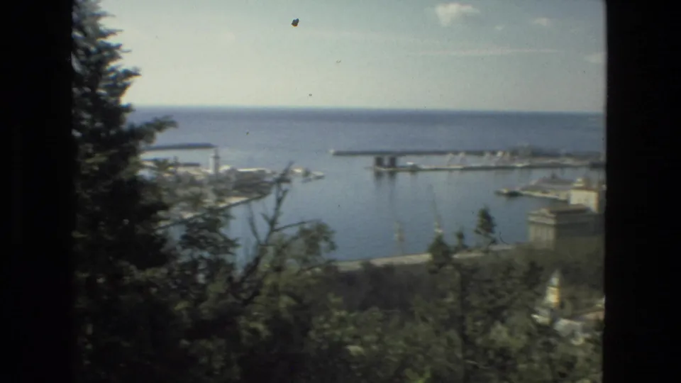 Elevated 1978 Spain travel footage frame showing harbor piers, open sea, trees, shoreline buildings, and small boats from a shaded overlook.