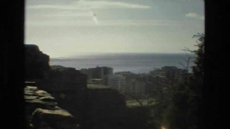 Soft 8mm travel frame showing a sea horizon, hillside or waterfront buildings, dark foreground stonework, and trees at the edge of the view.