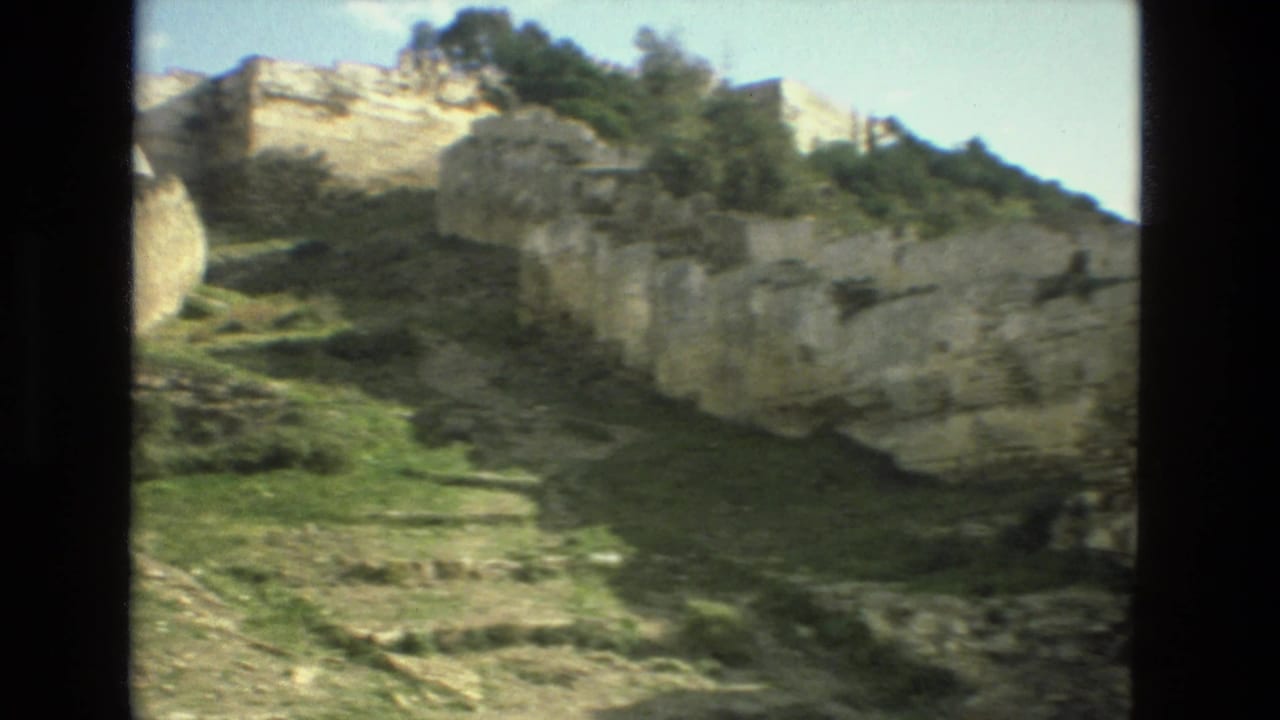 Stone ruins and pale rock on a grassy hillside under a bright sky.