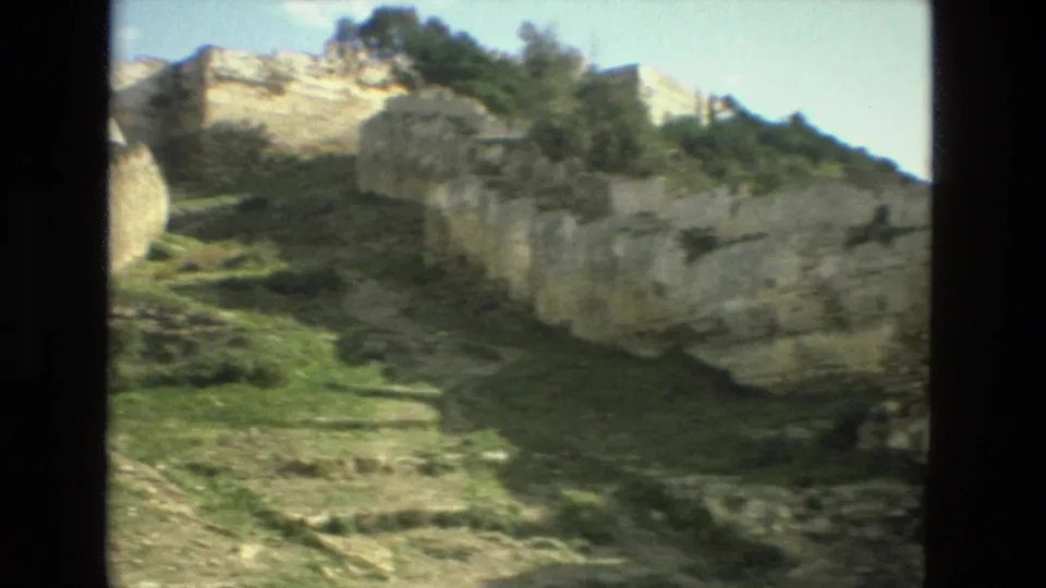 1978 8mm Granada view of weathered stone ruins, pale rock faces, grass, and trees on a sloping hillside.