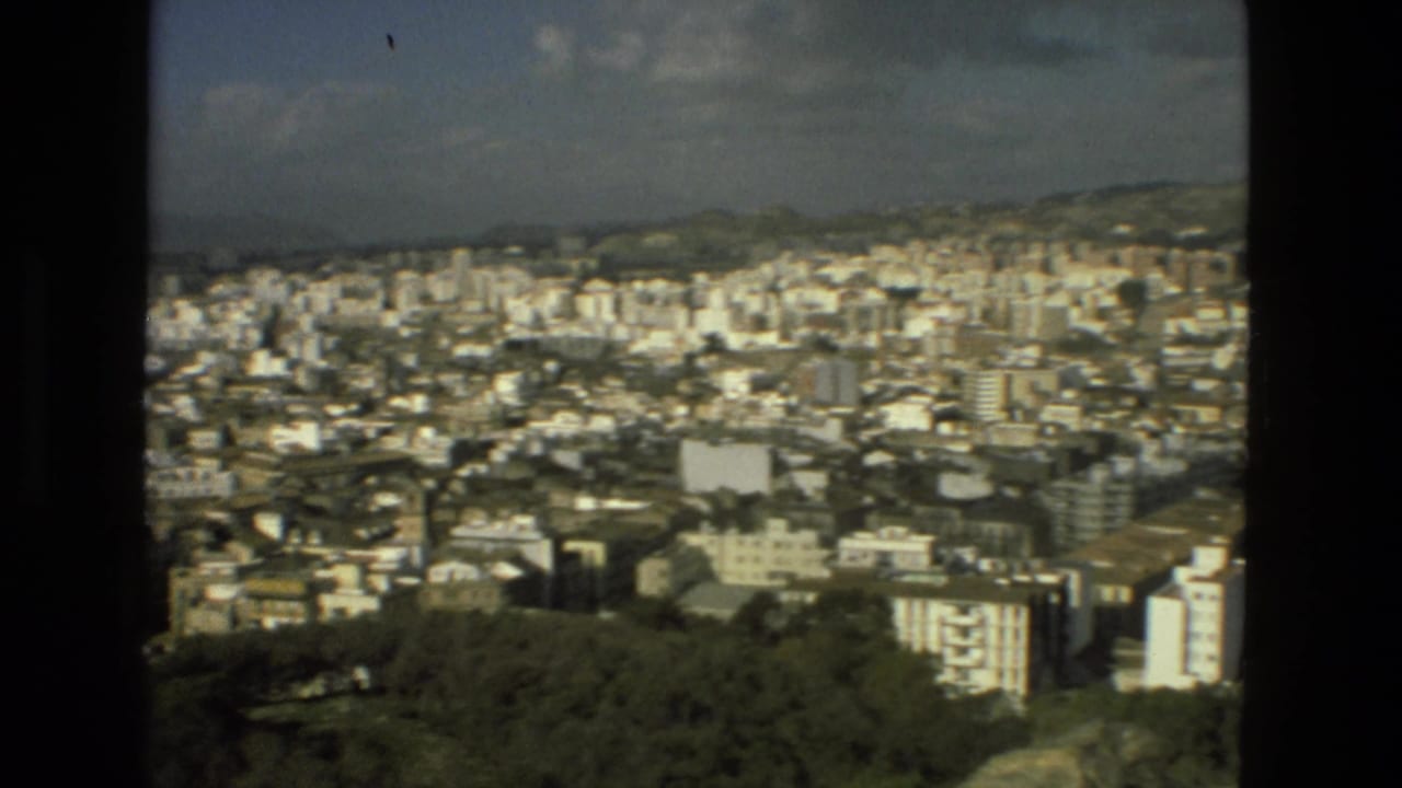 Wide elevated view over Granada with dense buildings, trees, distant hills, and dark film-frame borders.