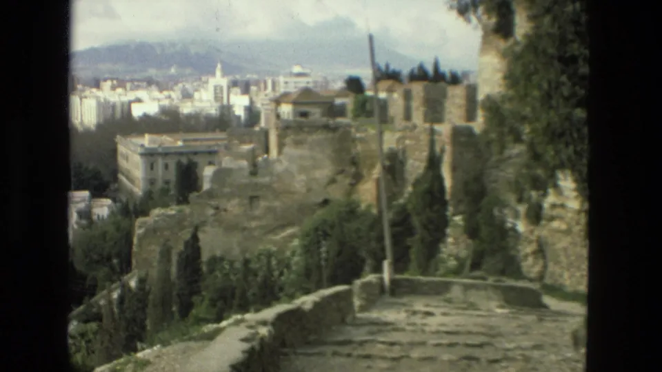 1978 8mm frame from Granada showing weathered stone walls, a path or steps, trees, urban blocks, and distant hills under cloud.
