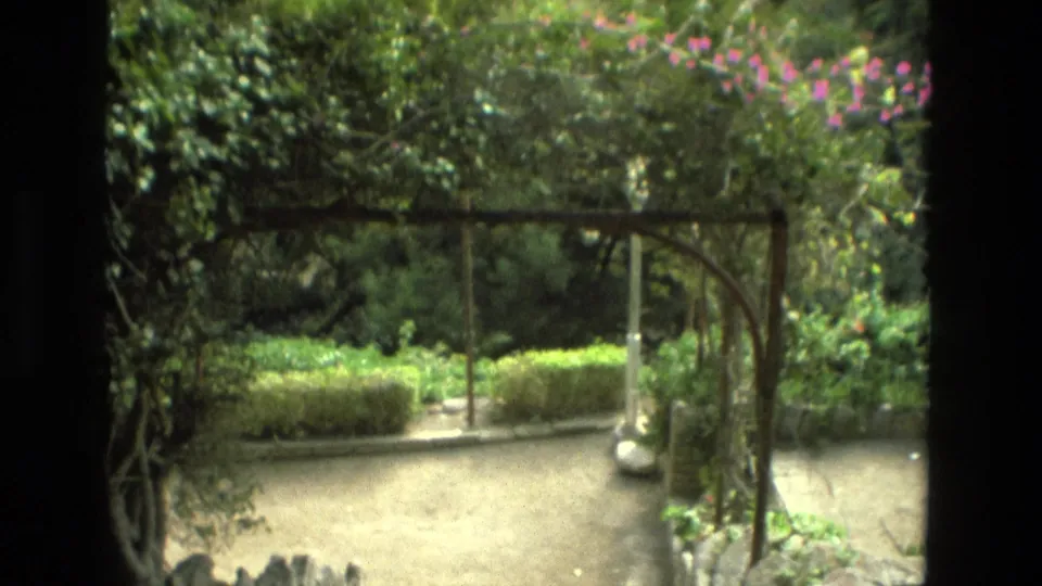 1978 Granada home-movie frame showing an arbor over a quiet garden courtyard with hedges and pale paths.