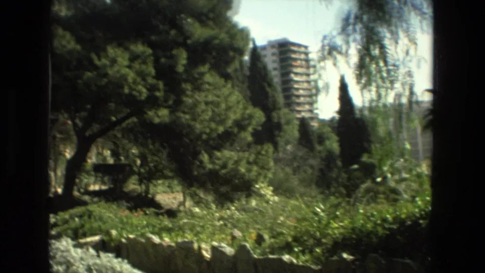 Supported Granada home-movie frame showing shaded trees, low garden planting, stone edging, and a distant apartment tower.