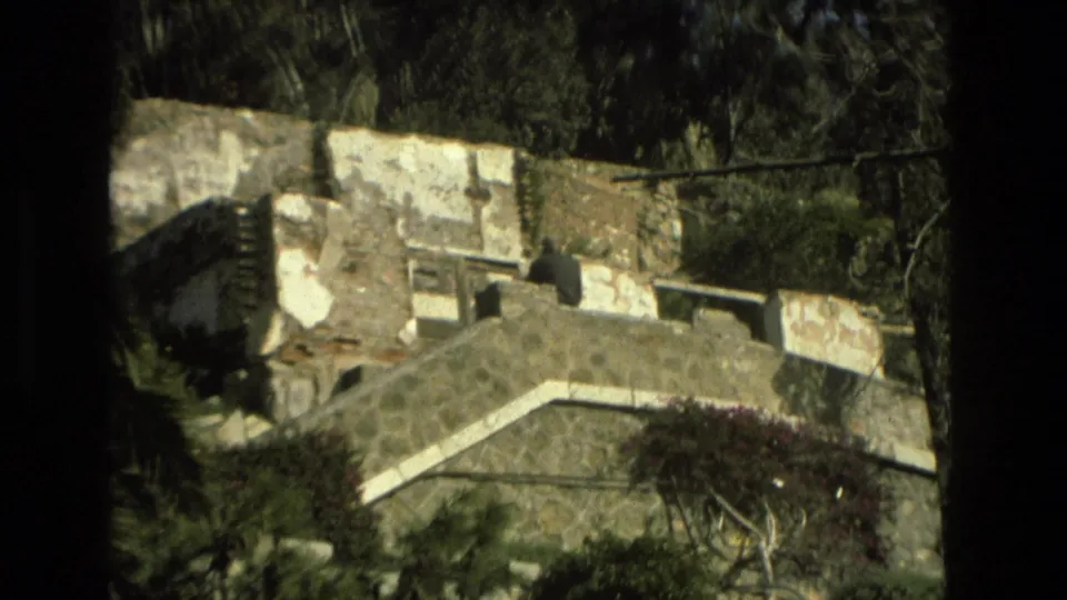 Granada home-movie frame with old stone stairs, rough masonry, greenery, flowers, and a distant person standing on the structure.