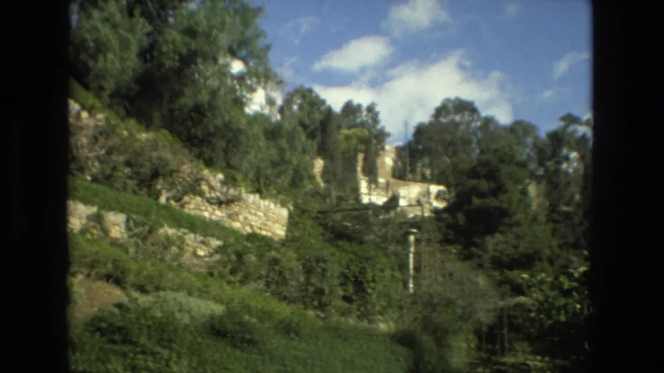 1978 Granada frame with a stone retaining wall, dense hillside foliage, tall trees, and pale ruin-like structures in the distance.