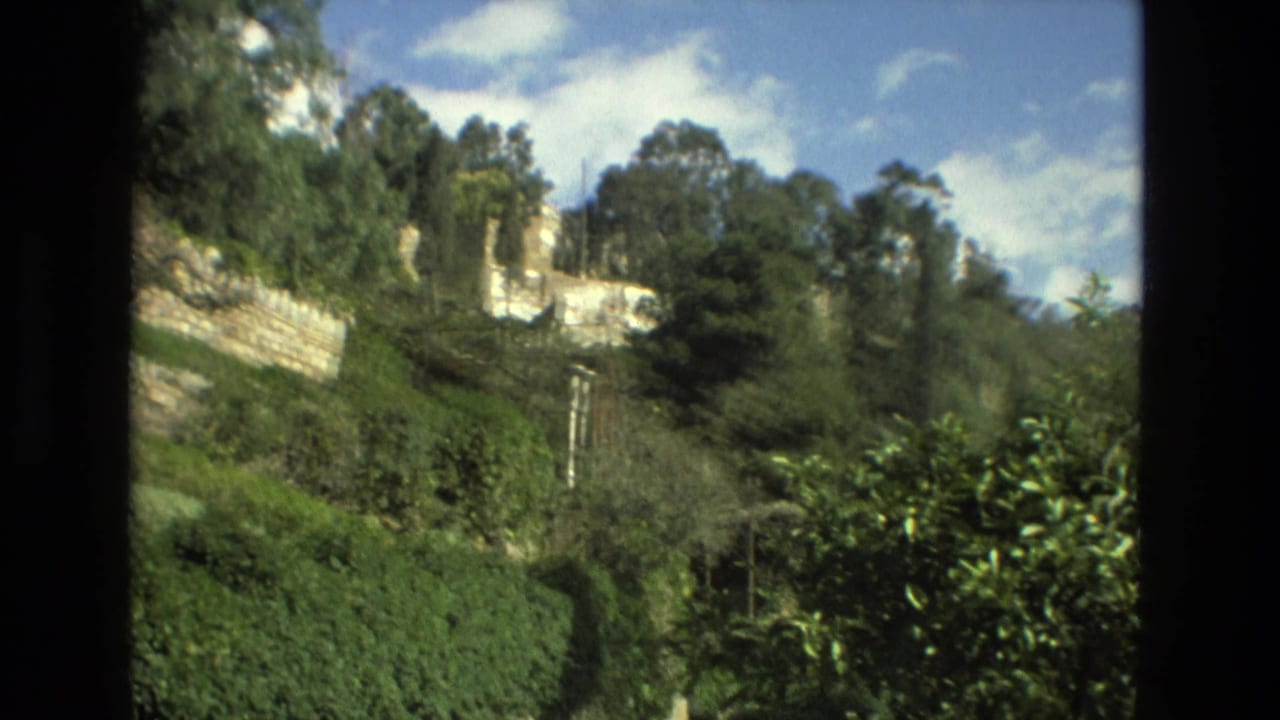 1978 Granada home-movie frame showing a green hillside garden, stone retaining walls, trees, sky, and distant buildings through a window-like divider.