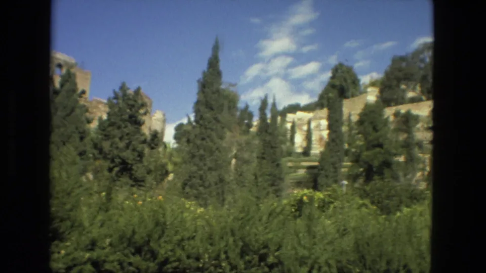 Granada home-movie view of sunlit stone ruins and tall cypress trees, with soft 8mm color and a clear sky.
