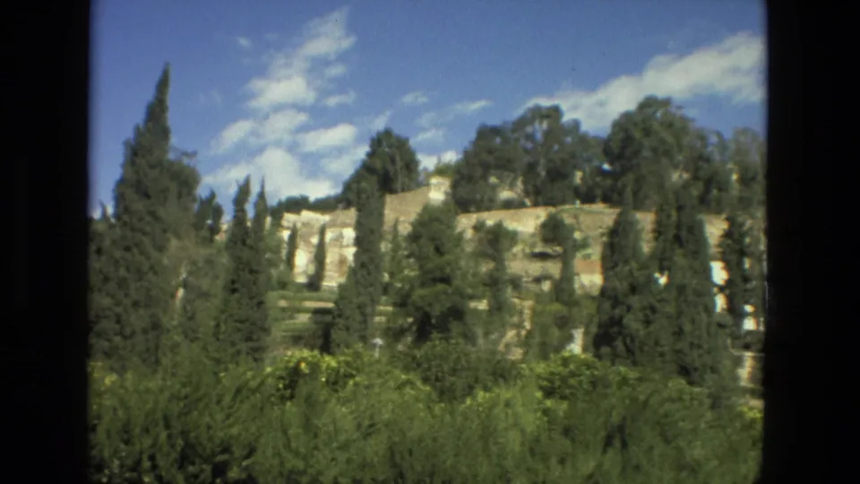 1978 Granada 8mm frame with cypress trees, dense foreground shrubs, and pale historic stonework beneath scattered clouds.