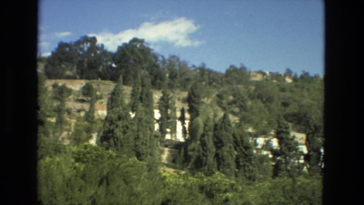Tree-covered hillside with pale cliff faces and white buildings under a blue sky.