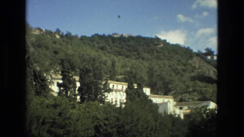 1978 Granada home-movie frame showing a tree-covered slope with white buildings partially hidden by foreground foliage.