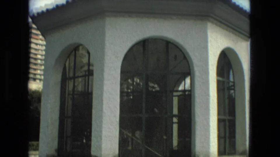 The home-movie camera moves closer across a white arched pavilion or gazebo in Granada, with glass panes, trees, and hillside buildings nearby.