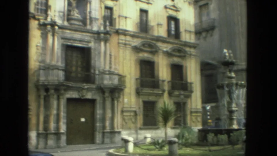 1978 Granada frame of an ornate stone facade with a large doorway, shuttered windows, balconies, fountain edge, and planted plaza area.