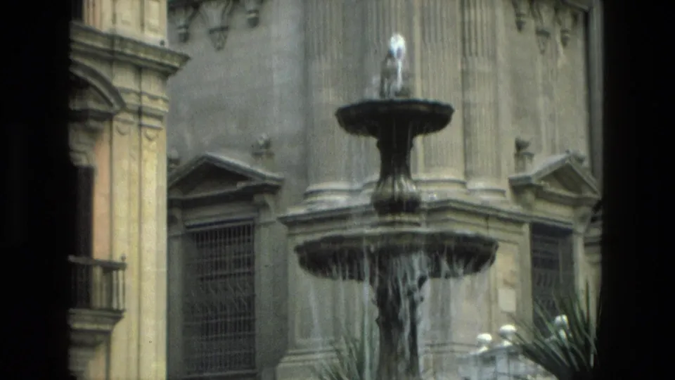 A Granada city-square fountain appears beside stone building facades, columns, barred windows, and planted palms in 1978 8mm footage.