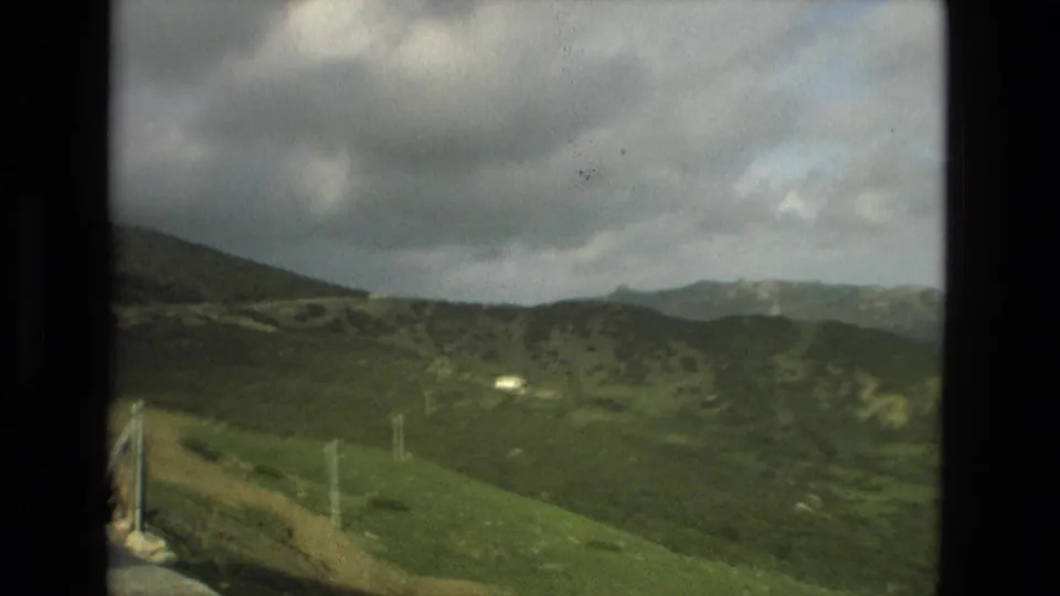 1978 8mm Granada hillside view with rolling green terrain, distant mountains, and small vertical posts near the foreground.