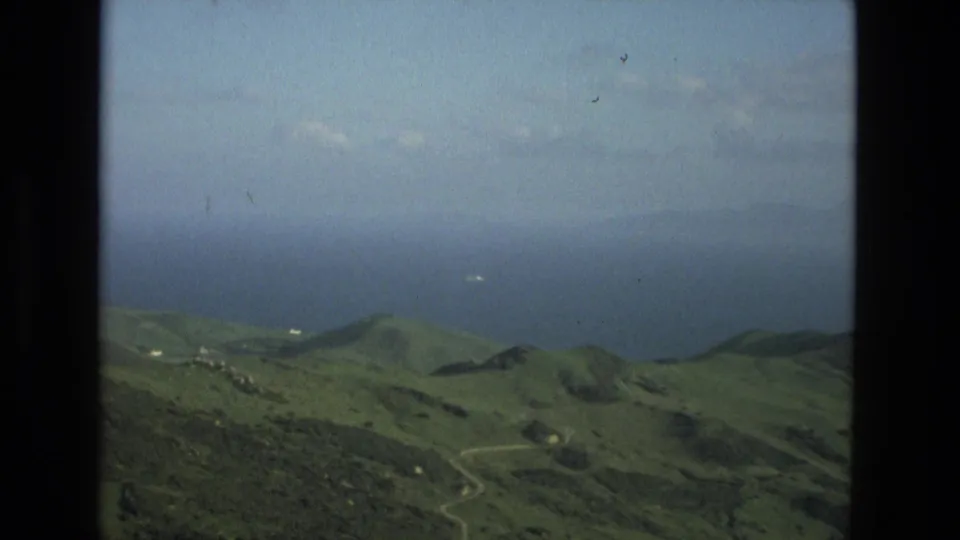 1978 Granada-area coastal mountain view with green hills descending toward blue water and a faint ship on the sea.