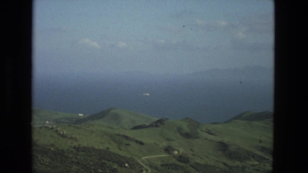 Green hills above distant blue water seen through a dark film-frame border.