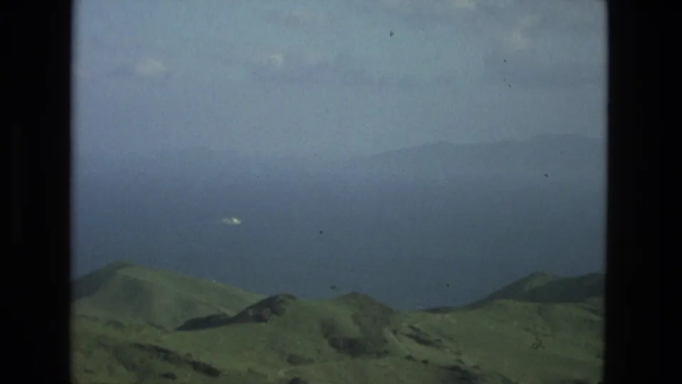 1978 8mm view from Granada, Spain area showing rounded green hills, blue water, haze, and a small white object in the distance.