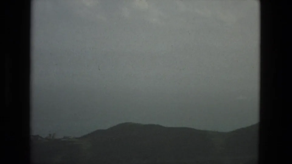 1978 Granada home-movie view of low dark hills under a pale grey cloud layer, with grain, dust, and the black edge of the film frame.