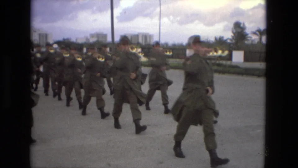 Uniformed soldiers in olive coats march in formation in Granada, Spain, with apartment blocks and cloudy sky in the background.