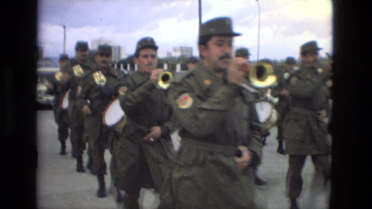 Uniformed military band members march on a paved street while playing brass instruments and drums.